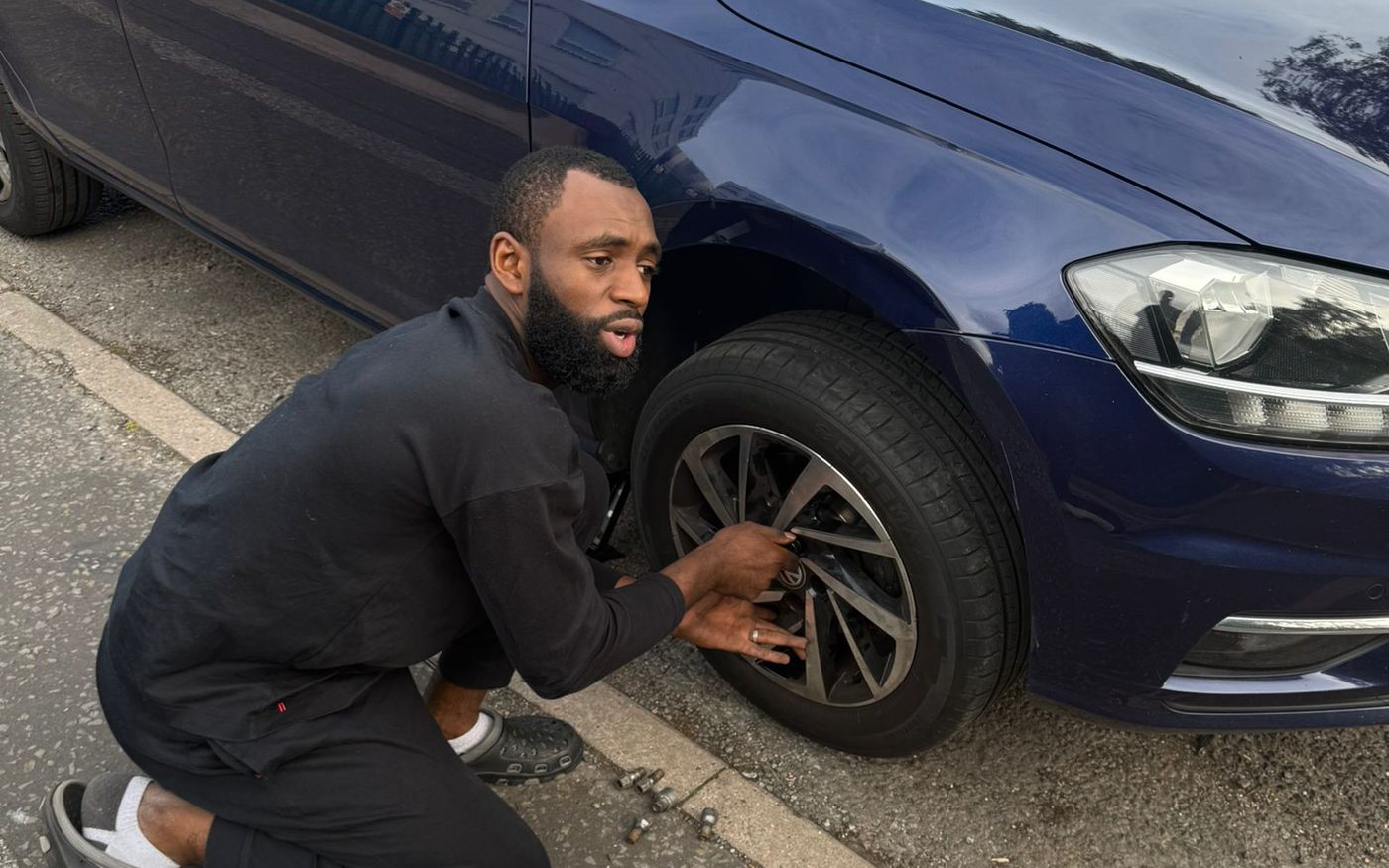 An Kerbside mechanic kneeling at the front wheel of a blue car at the kerbside